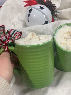 two green mugs filled with whipped cream on top of a white blanket next to a christmas ornament