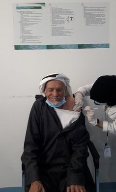 an old man sitting in a chair with another person wearing a mask on his face