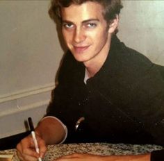 a man sitting at a table writing on a piece of paper with a pen in his hand