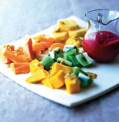 cut up fruit on a white plate with a red sauce in a glass pitcher next to it