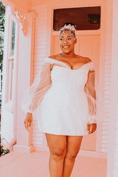 a woman in a short white dress standing on the front porch of a pink house