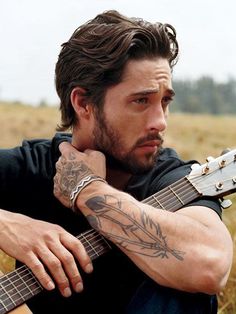 a man sitting in a field with an acoustic guitar and tattoos on his arm, looking at the camera