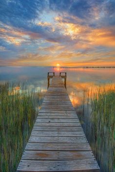 a wooden dock sitting on top of a lake under a colorful sky with clouds in the background