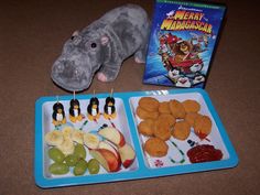 a toy hamster is sitting next to two trays of fruit and vegetables with an open book on the side