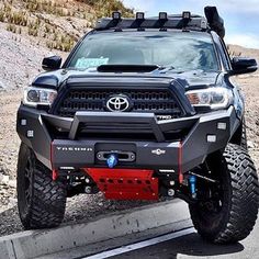 the front end of a black toyota truck parked on top of a mountain road with mountains in the background