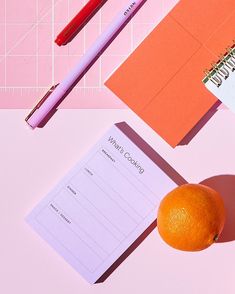 an orange sitting on top of a table next to a notepad and pencils