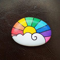 a painted rock with a rainbow sun and cloud on it's side, sitting on a brown leather surface