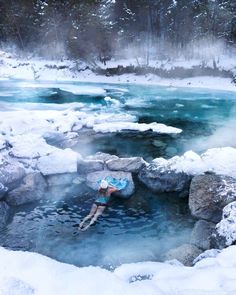 a person floating in a pool of water surrounded by rocks and snow covered ground with trees on either side