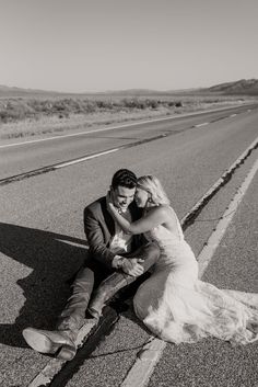 a bride and groom sitting on the side of an empty road in black and white