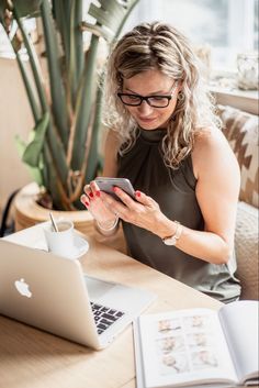 a woman sitting at a table looking at her cell phone and holding a laptop computer