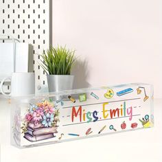 a clear box with an image of a book and flowers on the inside is sitting on a desk next to a potted plant