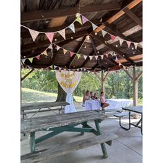 some people are sitting at a picnic table under a wooden structure with bunting and flags