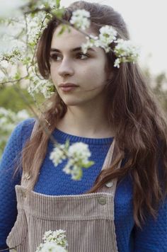 a young woman with long hair wearing an apron and holding flowers in her hand, looking at the camera