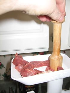 a person holding a wooden mallet over meat in a white dish on a counter