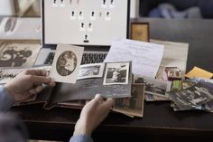 a person holding an open book in front of a laptop on a desk with papers and photos