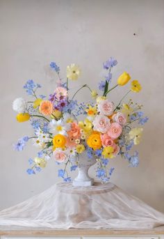 a white vase filled with lots of colorful flowers on top of a wooden table next to a wall