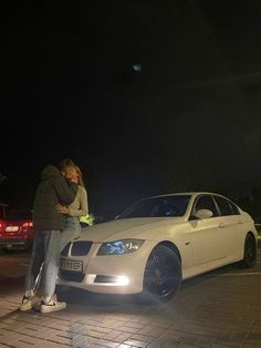 a man and woman standing next to a white car on the street at night with their arms around each other