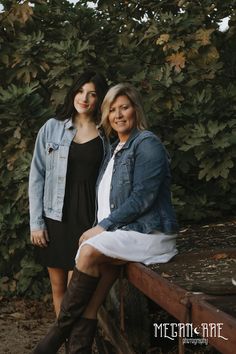 two women sitting on a wooden bench in front of some bushes and trees with their arms around each other