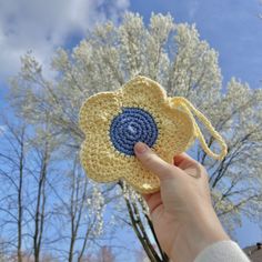 a hand holding up a crocheted flower in front of some trees and blue sky