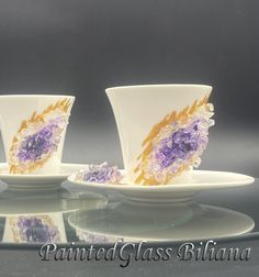 two white cups and saucers with purple flowers on them sitting on a glass table