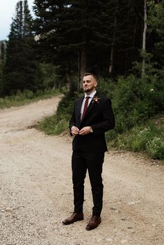 a man in a suit and tie standing on a dirt road next to some trees