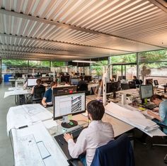 people working at desks in an open area with lots of windows and metal roofing