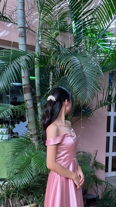 a woman in a pink dress standing next to some palm trees and looking up at the sky