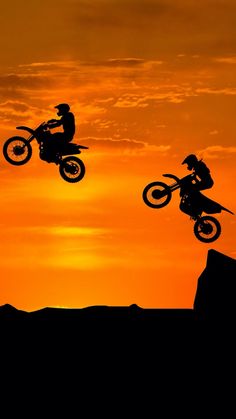 two people on dirt bikes jumping in the air at sunset or sunrise with one person doing a trick