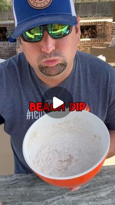 a man wearing sunglasses and a baseball cap holds a bowl of food in front of his face