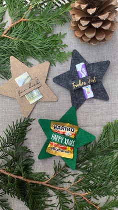 three felt star ornaments on a table next to pine cones and evergreen branches with the words happy xmas written on them