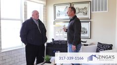 two men standing in a living room talking to each other and looking at something on the wall