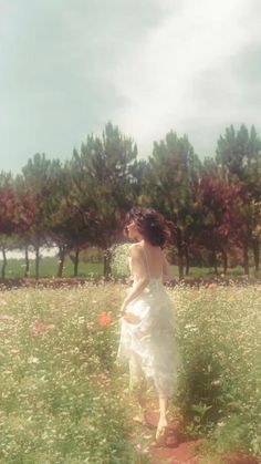 a woman in a white dress walking through a field with flowers and trees behind her