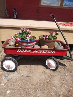 a red wagon filled with potted plants sitting on top of a dirt ground next to a house