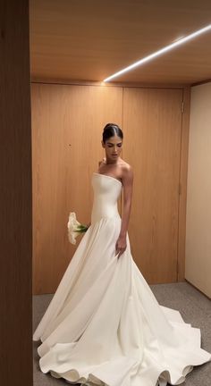 a woman in a white wedding dress standing next to a wooden wall and holding a flower