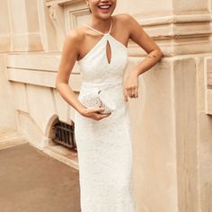 a woman in a white dress standing next to a wall holding a purse and smiling at the camera