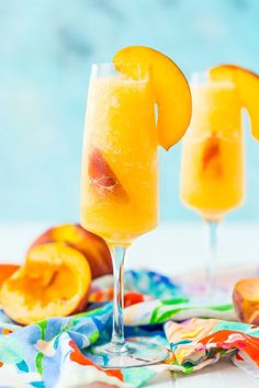 two glasses filled with orange juice on top of a colorful table cloth next to sliced peaches