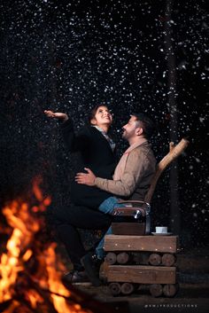 a man and woman sitting next to a fire in the dark with their arms around each other