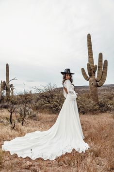 a woman in a long white dress and cowboy hat standing next to a large cactus