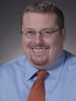 a man in a blue shirt and orange tie smiling at the camera while wearing glasses