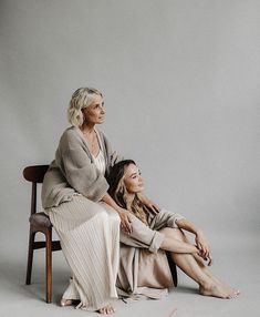 two women sitting next to each other on top of a wooden chair in front of a gray wall