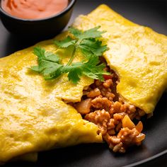 an omelet with meat and garnish on a black plate next to a small bowl of sauce