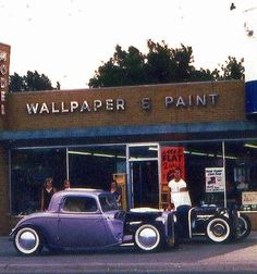 an old car parked in front of a wallpaper and paint shop with people standing outside