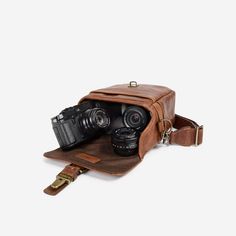 two cameras sitting in a brown leather bag on top of a white surface with a strap around it