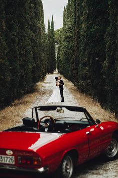 a bride and groom standing in front of a red convertible parked on the side of a road