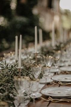 a long table is set with white plates and silverware, candles and greenery