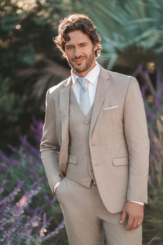 a man wearing a tan suit and tie standing in front of some purple flowers with his hands in his pockets