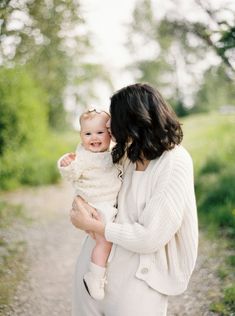 a woman holding a baby in her arms while standing on a dirt road next to trees