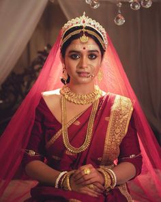 a woman wearing a red and gold bridal outfit sitting in front of a chandelier