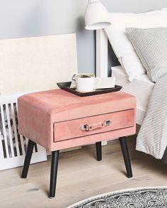 a pink table with a cup on it next to a white bed and black legs