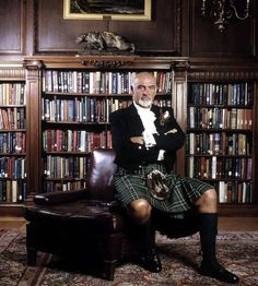 a man in a kilt sitting on a chair next to a book shelf filled with books